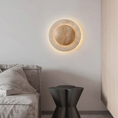 A modern round wall sconce made of natural stone with a wooden accent, illuminated by a warm LED halo effect, mounted on a white wall above a grey sofa.