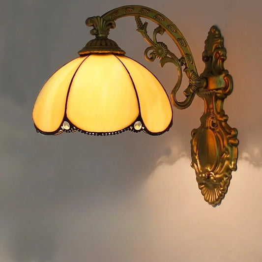 A wall-mounted light fixture with an antique gold metal arm and a scalloped yellow stained glass shade adorned with small crystals, illuminated against a dark background.
