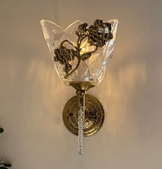 Close-up of a gold wall sconce featuring a clear glass shade with intricate geometric facets and a metallic floral embellishment, illuminated from within.