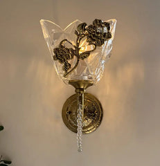 Close-up of a gold wall sconce featuring a clear glass shade with intricate geometric facets and a metallic floral embellishment, illuminated from within.