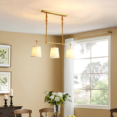 Polished copper linear pendant light with three white fabric shades hanging over a dining table.