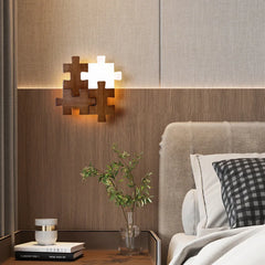 A wooden puzzle-shaped wall sconce with one illuminated white acrylic piece, mounted on a wood-paneled wall above a nightstand in a bedroom setting.