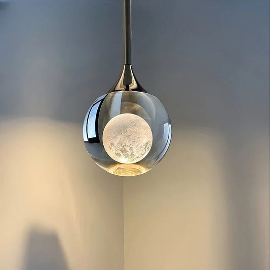 Clear glass globe pendant light with a textured inner orb resembling a moon, mounted on a brushed metal stem, casting light on a grey wall.