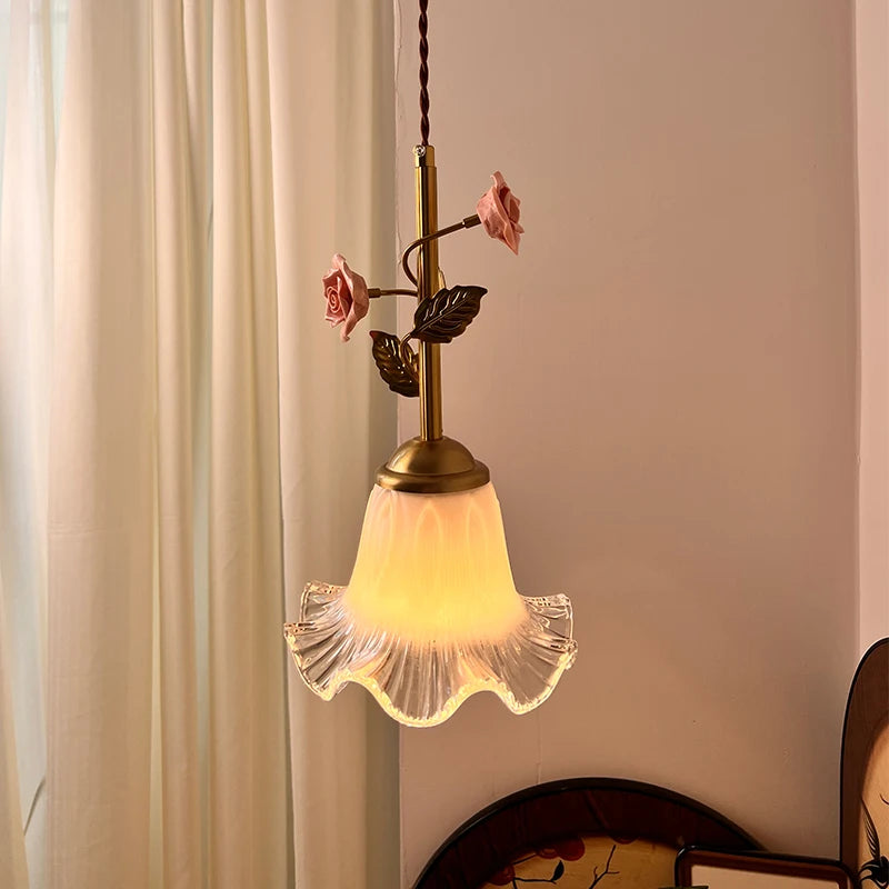 A pendant light with a clear fluted glass shade and a brass stem featuring pink ceramic roses and green leaves, illuminated warmly.