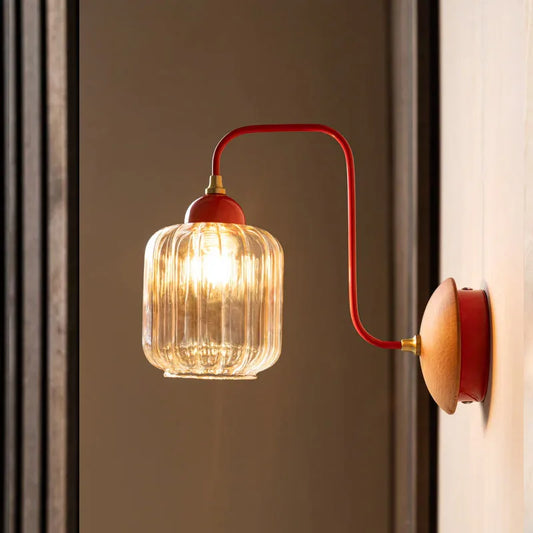 A wall sconce with a curved red arm and a clear ribbed amber glass shade illuminated by a warm LED bulb, mounted on a wood-paneled wall.
