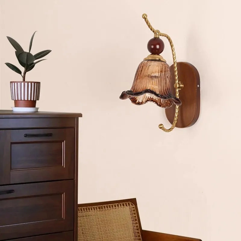 A vintage-style wall sconce with a floral-shaped amber glass shade and a gold arm mounted on a wooden oval backplate, shown in a living room setting.