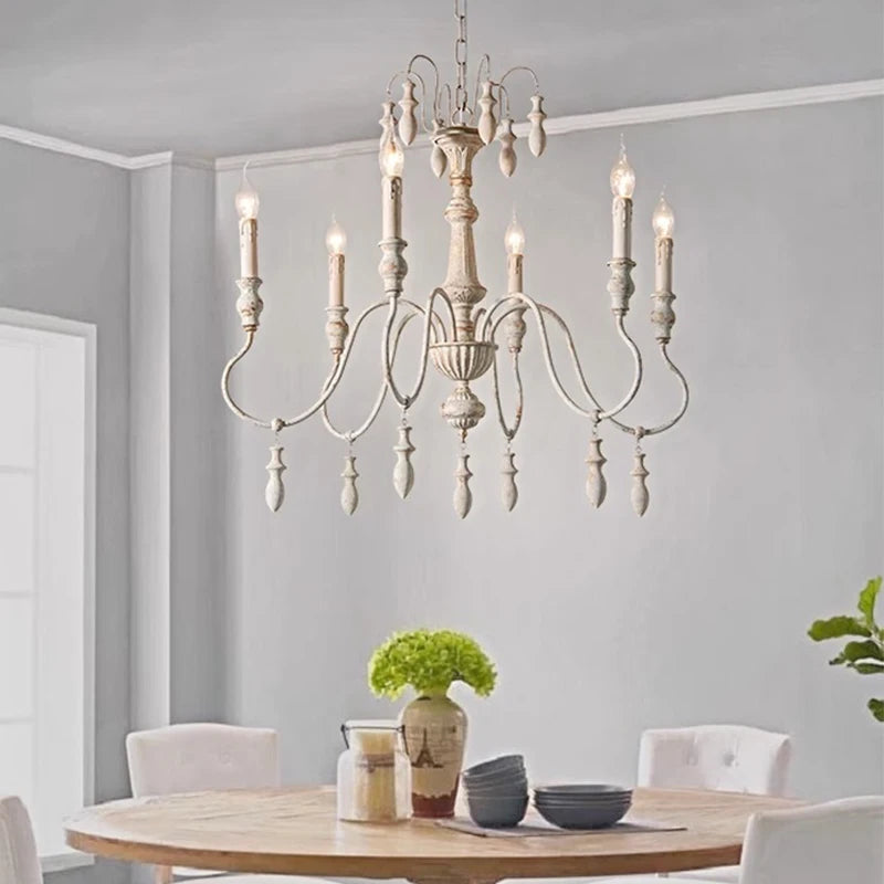 A distressed white iron candle-style chandelier installed in a dining room setting above a wooden table with chairs.