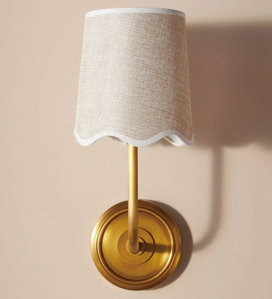 A wall sconce featuring a copper metal arm and base supporting a light-colored fabric shade with a scalloped trim, mounted on a pale pink wall.
