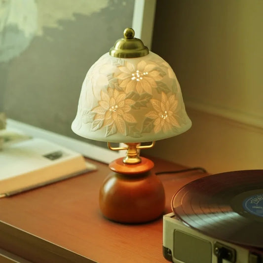 Table lamp with a white ceramic dome shade featuring embossed floral patterns and a brown wooden base, placed on a wooden surface next to a record player.