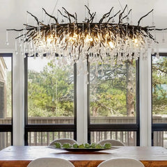 A modern black branch-style chandelier with numerous teardrop crystals hanging above a wooden dining table with a scenic forest view through large windows.