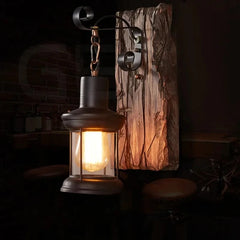 A black iron lantern wall sconce with a distressed wood backplate, casting a warm glow in a dimly lit room with brick accents.