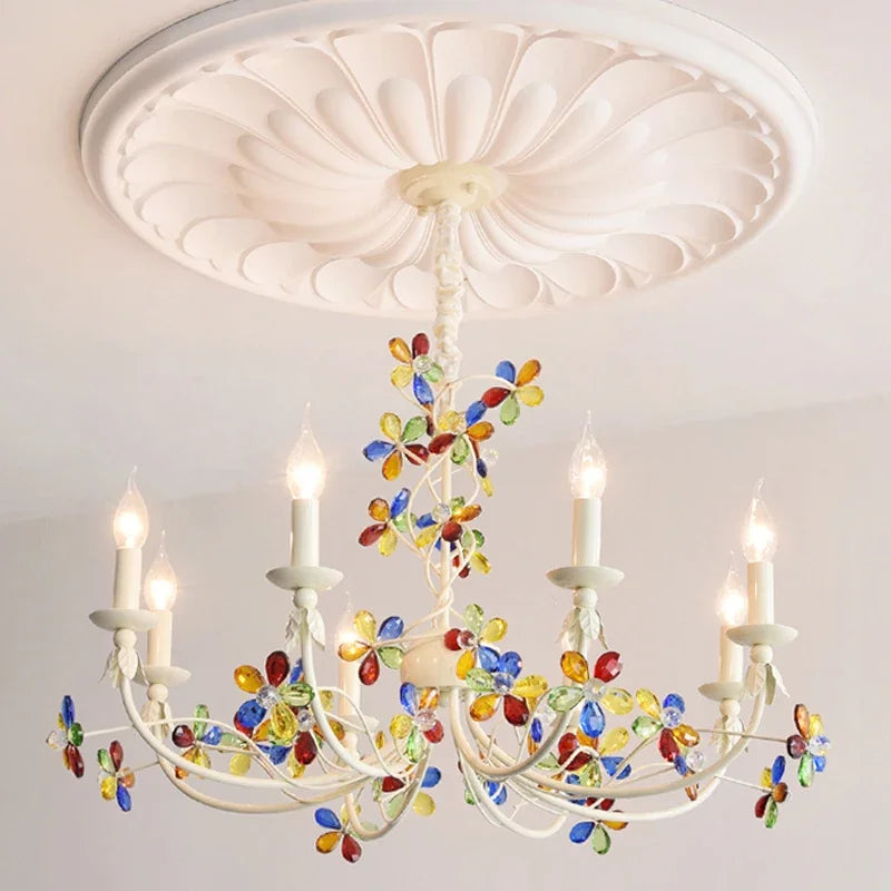 A white candelabra-style chandelier featuring numerous colorful crystal flower embellishments and lit candle bulbs.
