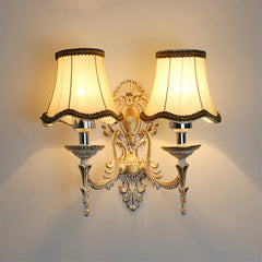 A dual-light gold wall sconce with cream fabric shades, displaying an ornate central backplate and curving arms.