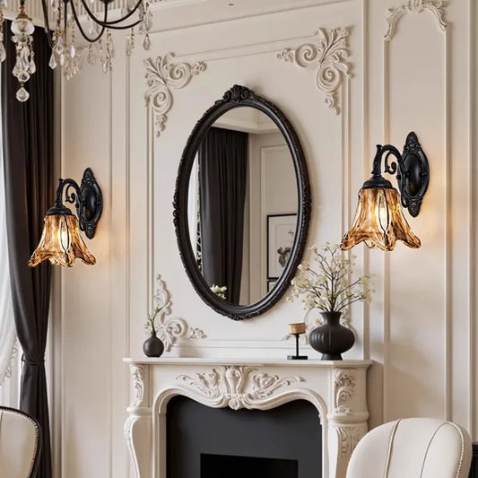 Two bronze glass bell wall sconces flanking an oval mirror above a fireplace, in a richly decorated room.