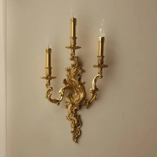 Gold candelabra-style wall sconce with three candle-shaped lights mounted on an ornate base against a light-colored wall.