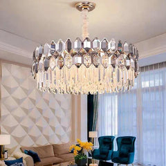 A round chandelier with multiple layers of faceted crystals, suspended by a cream cord from a white ceiling in a living room setting with modern furniture and patterned wallpaper.