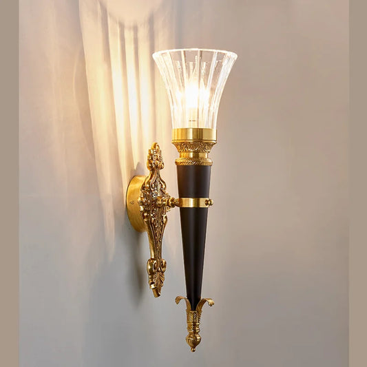 A gold wall sconce with a flared clear glass shade and a dark conical body, mounted on a gray wall, casting light patterns.