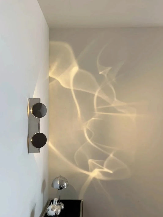 Close-up of a modern wall sconce with two dark circular discs on a silver mount, showing the light projection.