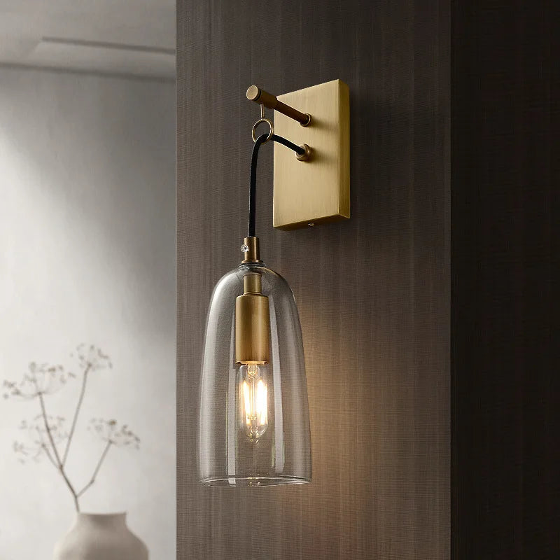 A modern gold and clear glass bell wall sconce mounted on a dark textured wall, with subtle lighting.