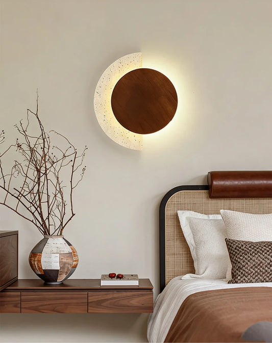 Modern crescent-shaped wall sconce with a wood circle and speckled stone accent, casting warm light on a bedroom wall above a nightstand.