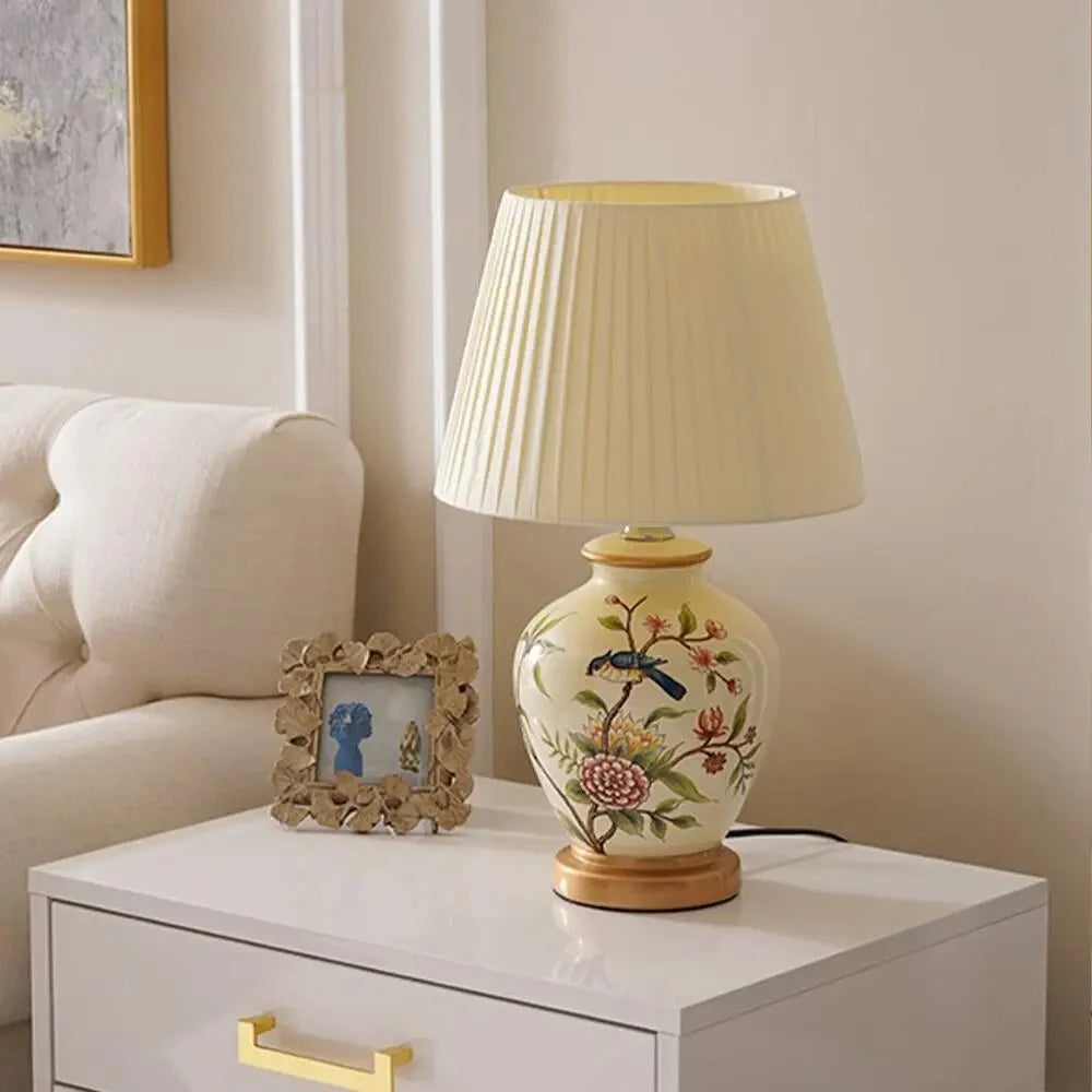 A ceramic table lamp with a white pleated shade, featuring a hand-painted bird on a floral branch, positioned on a white drawer unit next to a bed.