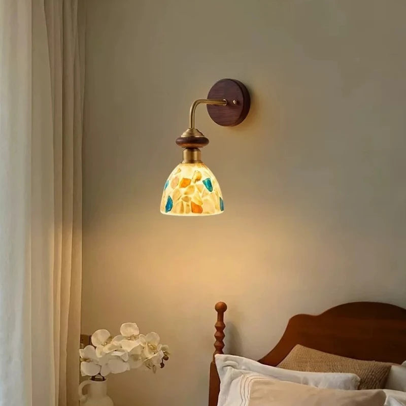 Wall sconce with a bell-shaped shade made of colorful shell fragments mounted on a wood base with a brushed gold arm.
