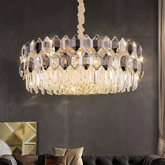 A multi-tiered chandelier with numerous faceted crystals in clear and smoky tones, suspended by a cream-colored cord from a white ceiling. The fixture has a gold-toned metal frame.