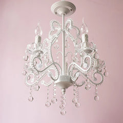 A white candelabra chandelier featuring crystal droplets hanging from its arms, illuminated against a pink wall.