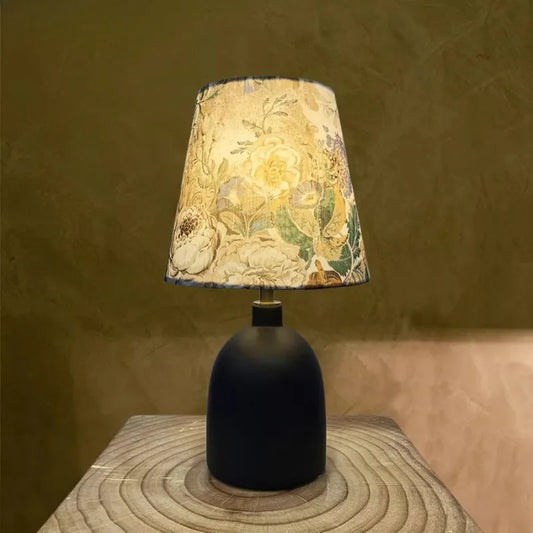 Close-up of a black ceramic table lamp with a floral fabric shade, illuminated in a warm setting against a textured wall.