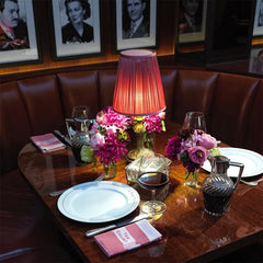 A table set for dinner with place settings, flowers, and a pinkish-red pleated fabric shade table lamp as a centerpiece.