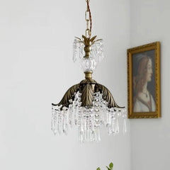 A brass crystal chandelier with a leaf-shaped canopy, softly lit and displayed in a room setting with a painting.