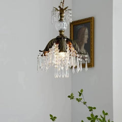 Brass crystal chandelier with a leaf-shaped canopy, illuminated and hanging in a room with artwork on the wall.