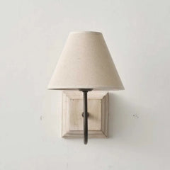 A wall sconce with a light beige fabric cone shade mounted on a distressed white square wood base, against a white wall. The fixture has a dark metal arm extending from the base to the shade.