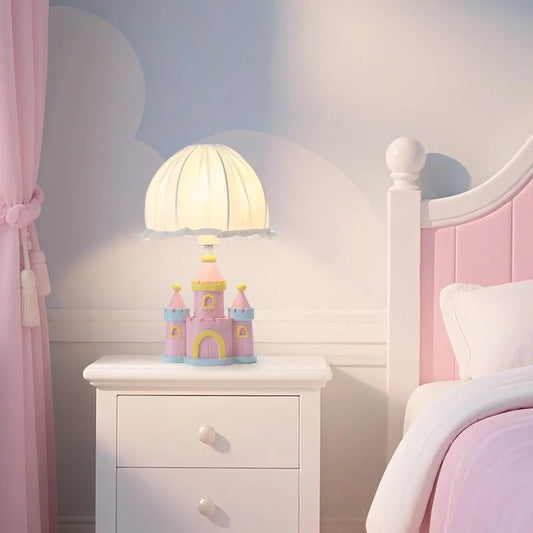 A pink castle table lamp with a pleated white fabric shade sits on a white bedside table next to a pink-tufted bed, with pink curtains visible.