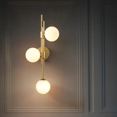 A modern gold wall sconce with three white frosted glass globes mounted vertically on a brass stem against a dark gray paneled wall.