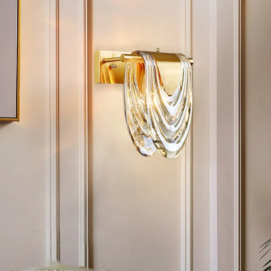 A gold wall sconce with a wavy clear glass shade, illuminated on a panelled wall.