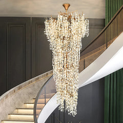 A dramatic gold crystal chandelier hangs in a modern foyer with dark wood paneling and a curved staircase featuring subtle lighting.