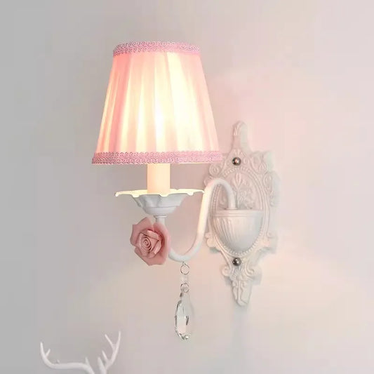 Close-up of a single white wall sconce with a pink pleated fabric shade, pink rose accent, and crystal droplet.