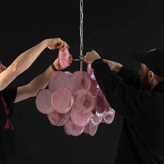 Two individuals assembling a pink glass disc chandelier against a black background.
