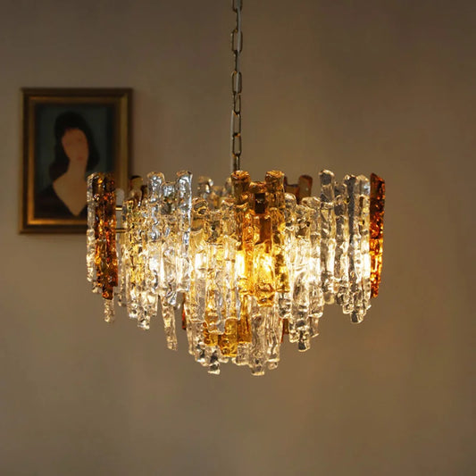 Close-up of a glass tube chandelier featuring a mix of amber and clear glass tubes illuminated by internal lights, with a framed portrait in the background.