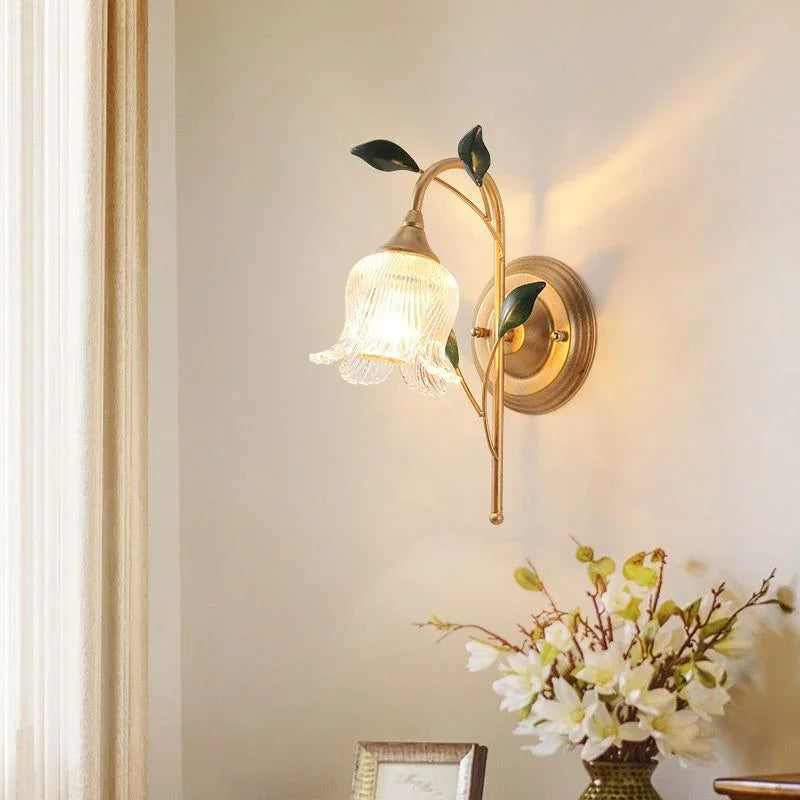 Gold flower wall sconce with clear glass shade in a room setting with white walls, a cream curtain, and a vase of flowers on a table below.