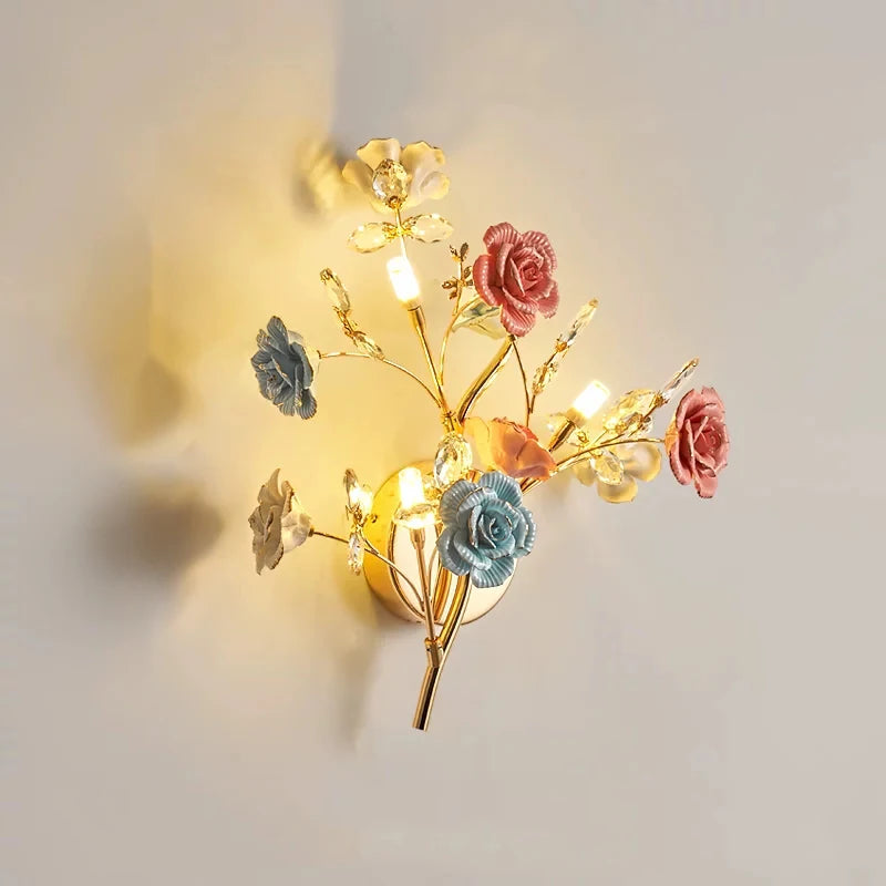 A gold wall sconce with a mix of pink, blue, and white ceramic flowers and clear crystals, emitting a warm light.