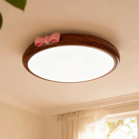 A round, flush mount ceiling light with a wood-tone frame and a pink bow accent, emitting a soft white light against a white ceiling.
