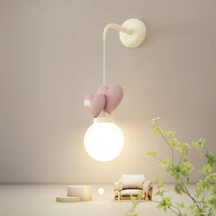 A wall sconce with a cream mounting bracket, a white cord, three pink heart-shaped accents, and a white globe light emitting warm light.