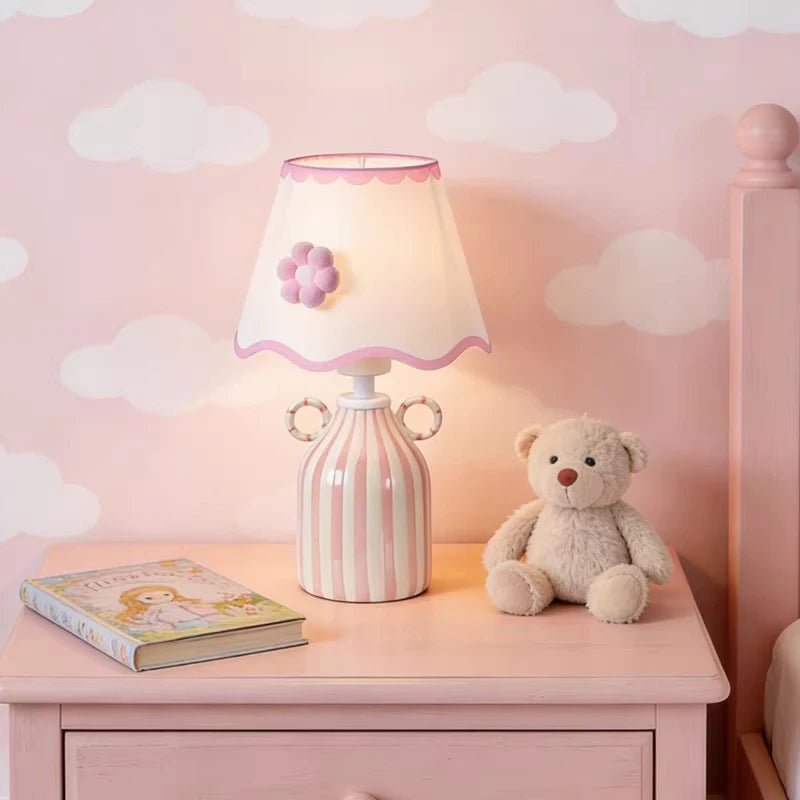 Pink and white striped ceramic table lamp with a scalloped white fabric shade featuring a pink flower accent, placed on a pink nightstand next to a teddy bear and a book.