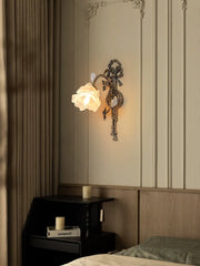 Close-up of an antique gold flower wall sconce with a white frosted glass shade, mounted on a wall with decorative paneling and a black nightstand beside it.