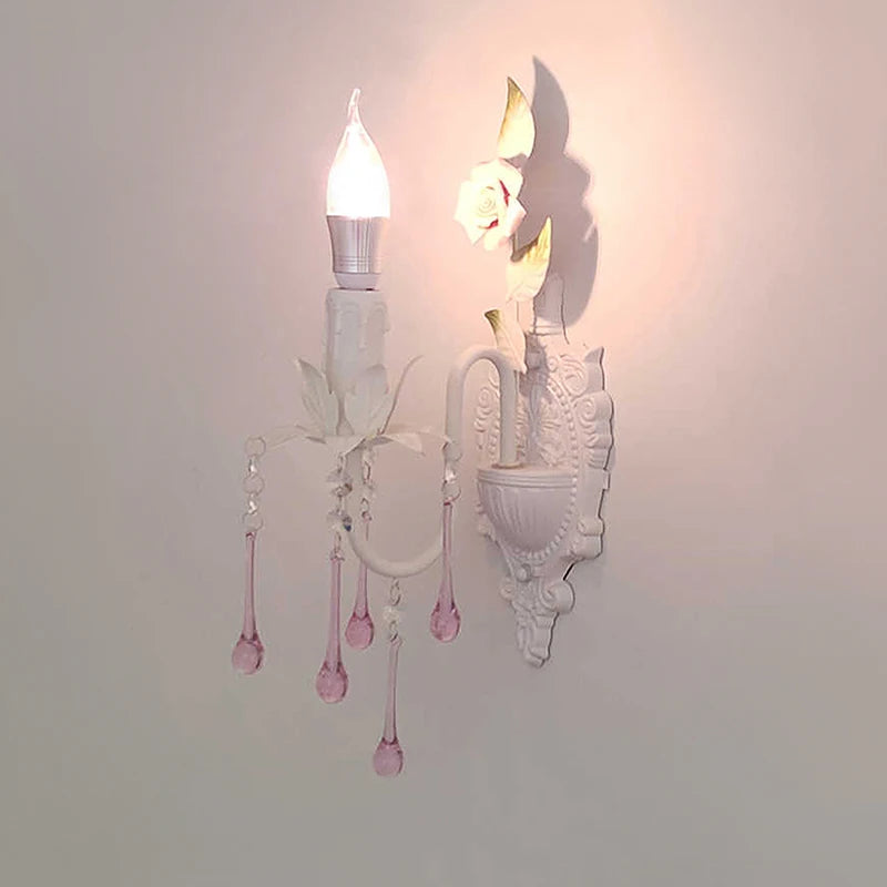 A lit white wall sconce with pink crystal droplets, casting a shadow on the wall, showcasing its floral design.