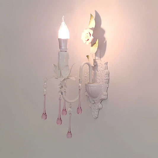 A lit white wall sconce with pink crystal droplets, casting a shadow on the wall, showcasing its floral design.