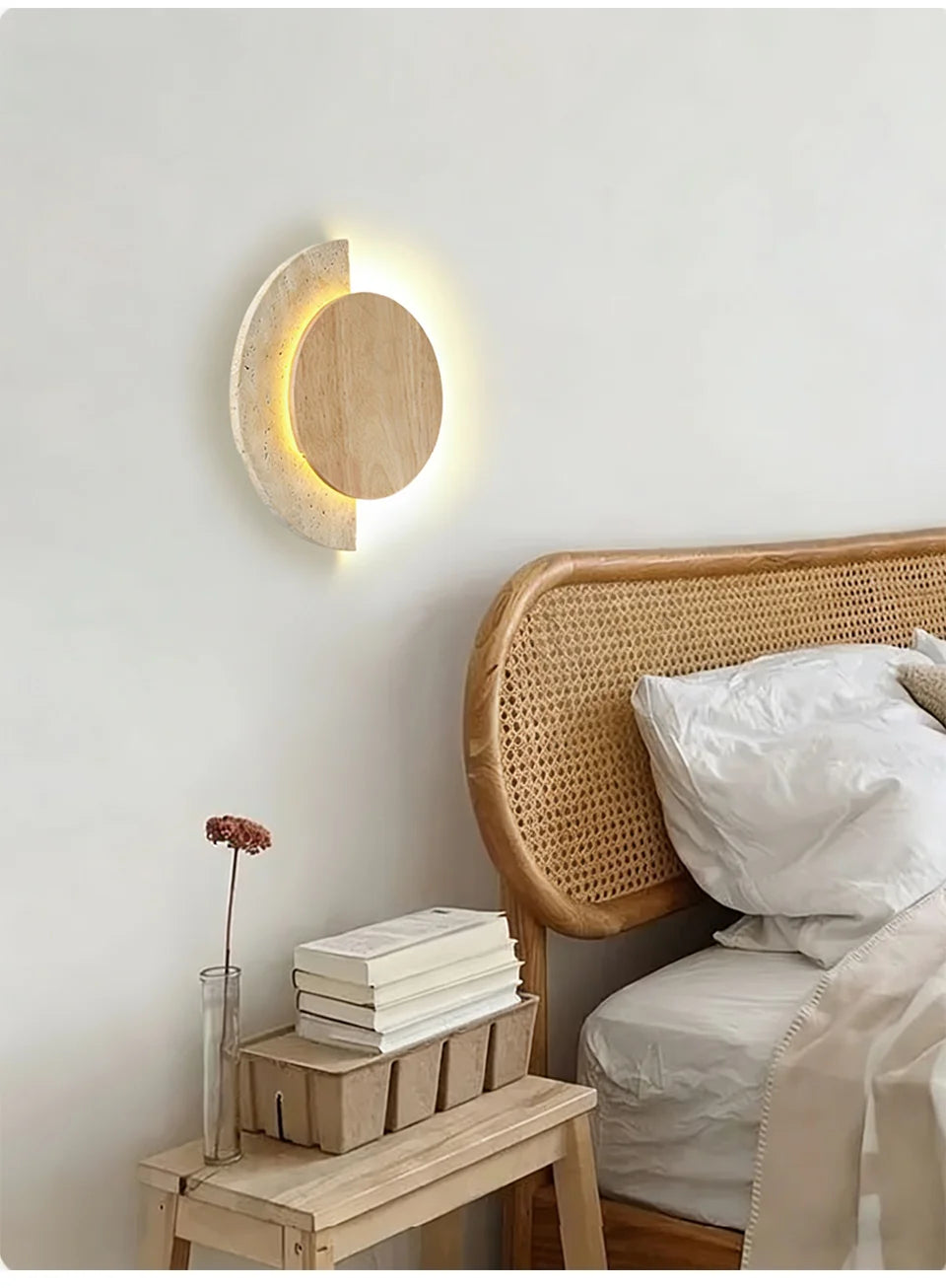 A modern wall sconce with a wood circle and speckled stone arc mounted on a wall above a bedside table with books.