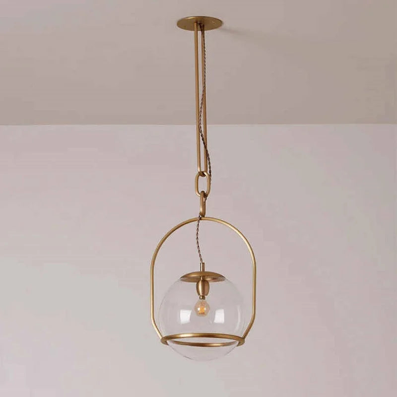 A gold metal frame surrounds a clear glass globe pendant light, suspended by a chain from a ceiling mount.